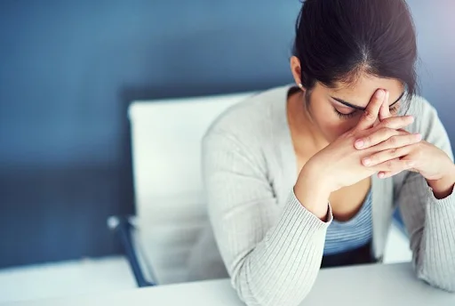 A woman contemplates with her head bent while slightly pinching her nose and forehead while on a work desk.