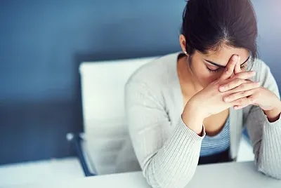 A woman contemplates with her head bent while slightly pinching her nose and forehead while on a work desk.