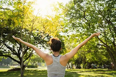 A woman stands in a sunlit park with her arms raised.