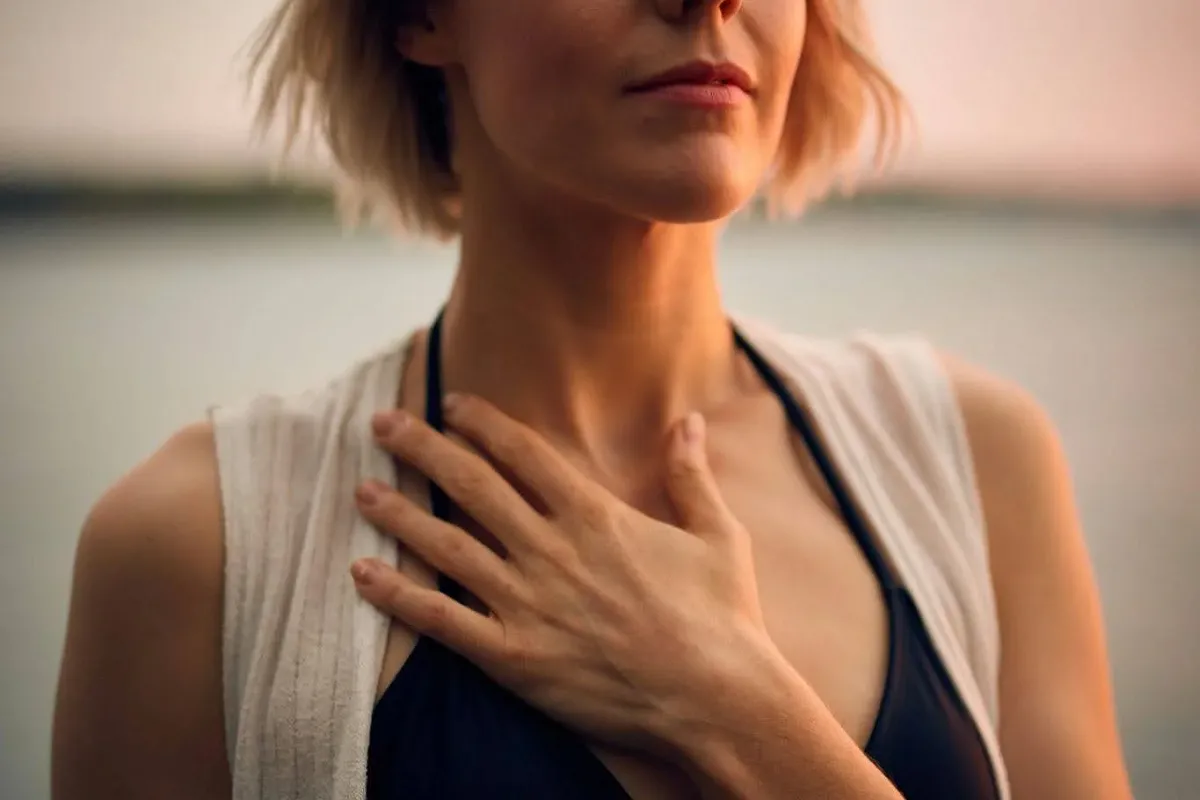 A woman stands in a sunlit park with her arms raised.